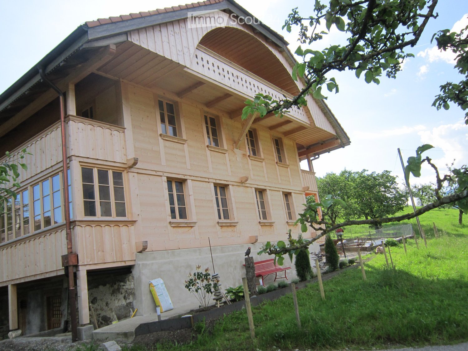 House. Two-story wooden house with balconies, large windows, and a stone base. The house is surrounded by greenery and a small garden area with a red bench.