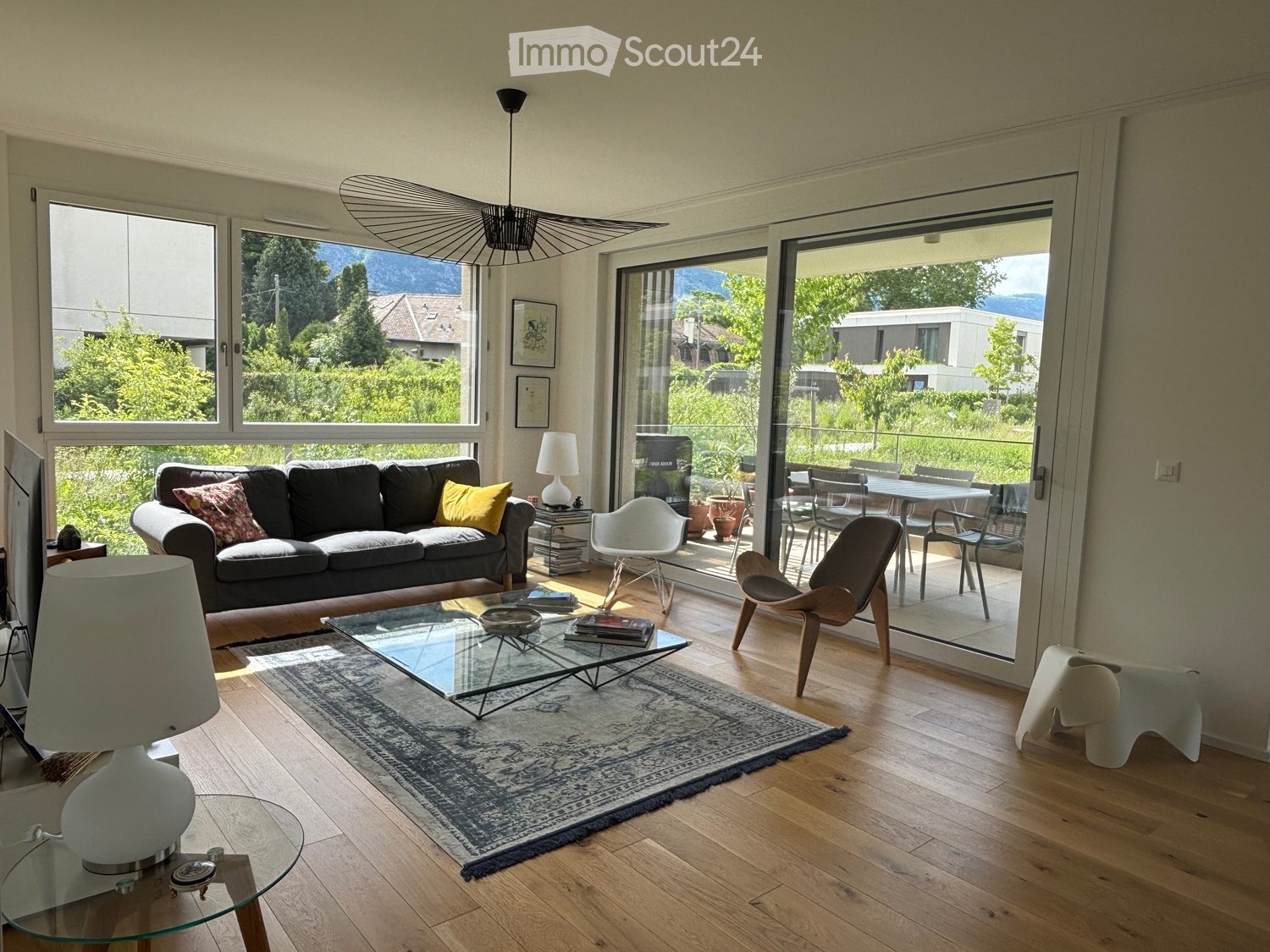 Living Room. Modern living room with large windows and a view, featuring a dark grey sofa, glass coffee table, white lamp, and a dining area with chairs and a table visible through the open sliding doors.