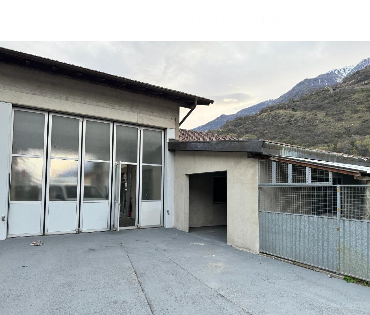 Building. Exterior view of a modern building with large glass doors, a covered entrance area, and a metal fence, set against a mountainous backdrop.