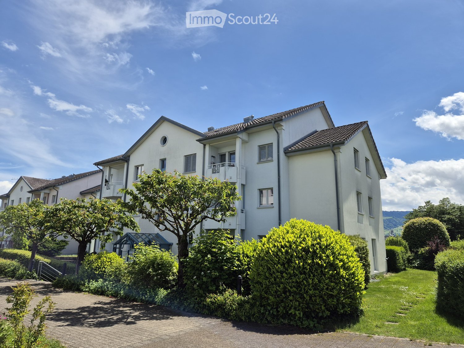 House. Modern two-story house with white exterior, multiple windows, and a balcony on the upper floor. The house is surrounded by well-maintained greenery, including trees and shrubs, with a paved driveway leading to the entrance.