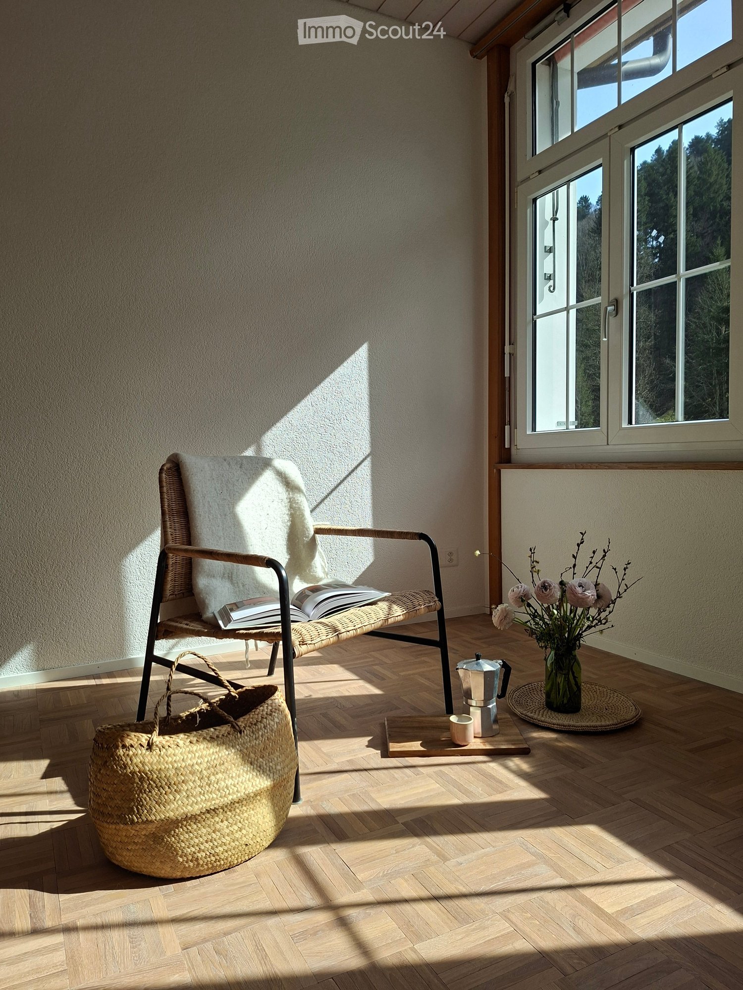 Furnished Room. A cozy, sunlit room with a wicker chair, a woven basket, a coffee maker, a cup, and a vase with flowers on a wooden floor.