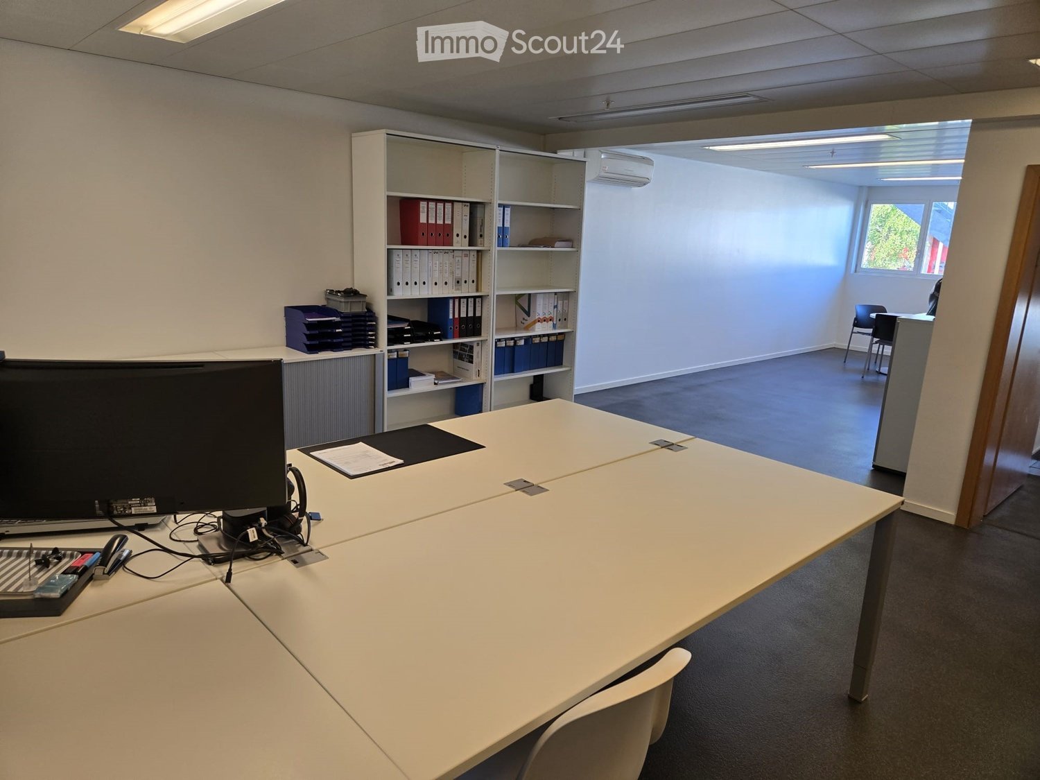 Furnished Room. Office space with a large white table, chairs, a screen, laptop, keyboard, mouse, and shelves with folders and books.