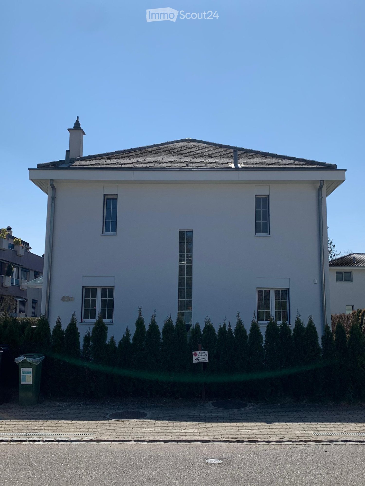 House. Two-story house with white exterior, multiple windows, and a chimney. The house is surrounded by neatly trimmed evergreen trees and has a paved driveway.