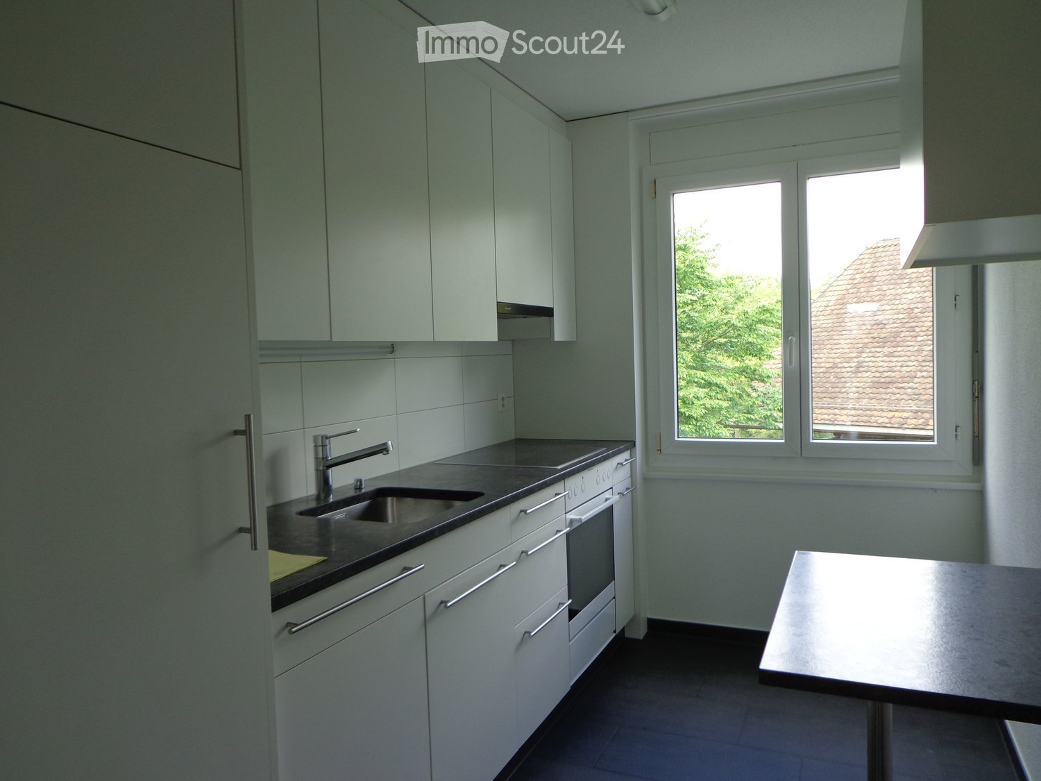Kitchen. Modern kitchen with white cabinets, stainless steel sink, faucet, and built-in oven. A small table is positioned near the window, which offers a view of the roof and greenery outside.