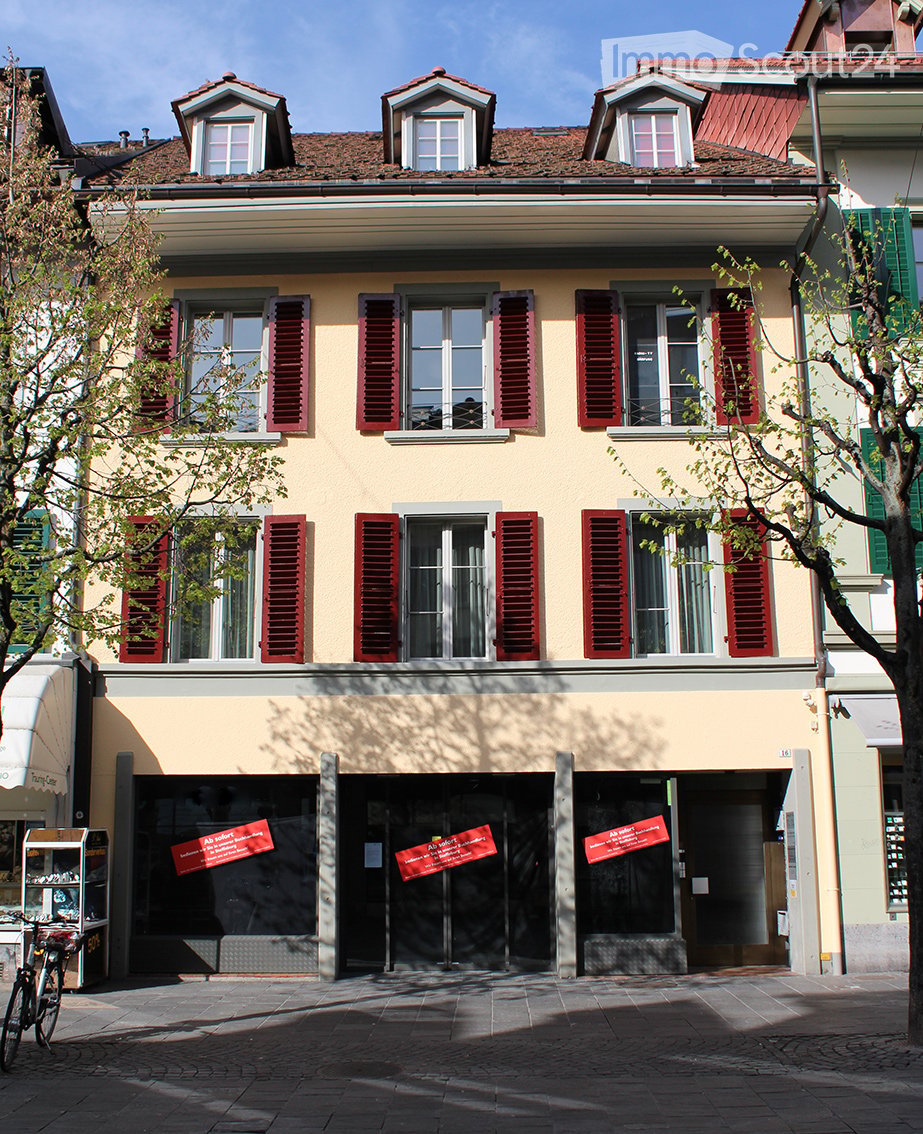 Three-story building with yellow facade, red shutters, and multiple windows. The ground floor has large glass doors with red banners, and there is a bicycle parked to the left.