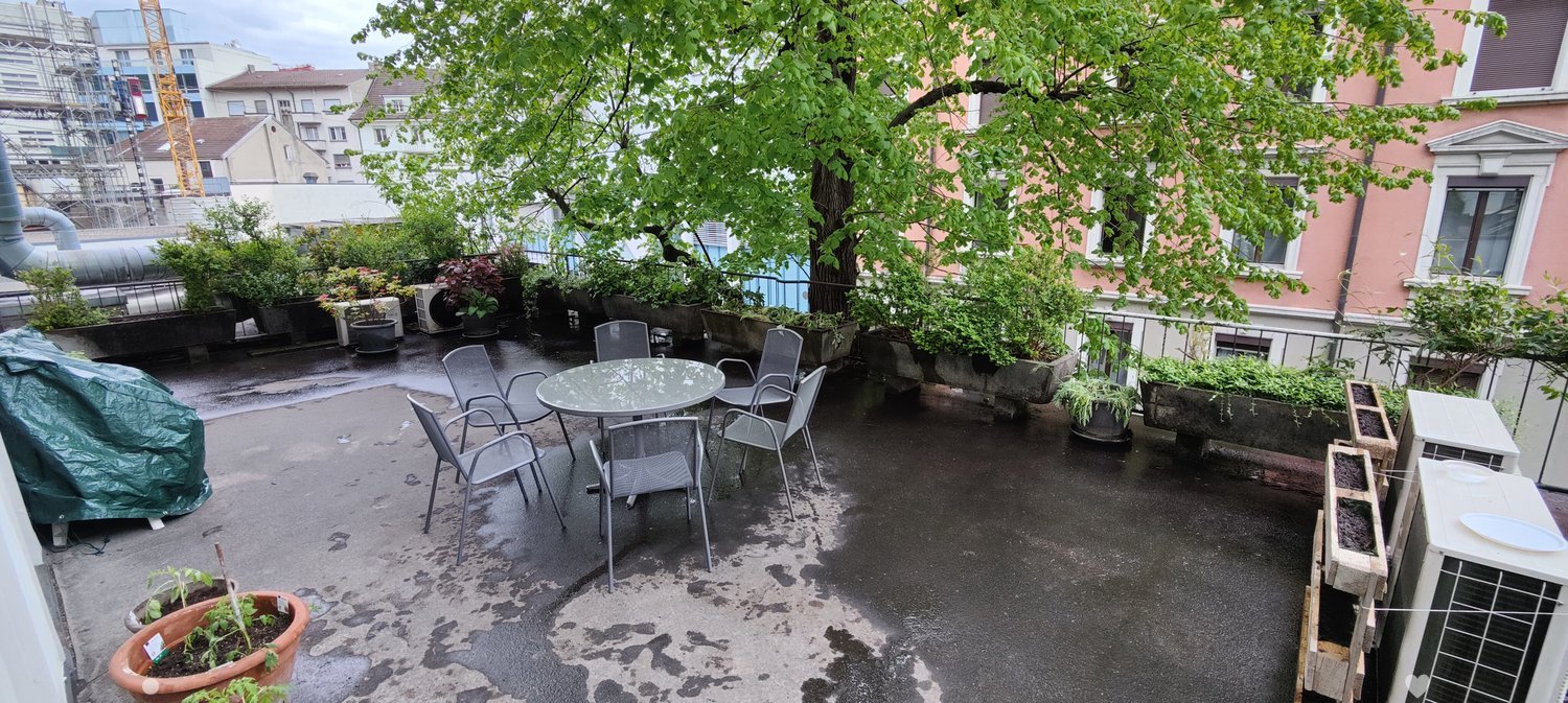 Balcony / Terrace. Balcony with a round table and chairs, surrounded by potted plants and trees, with a cityscape view in the background.