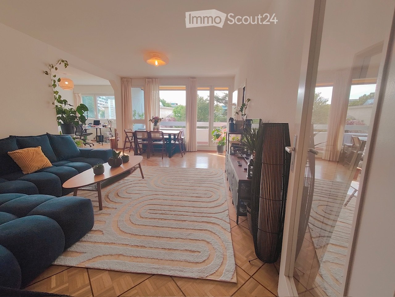 Furnished Room. Spacious living room with a blue sectional sofa, coffee table, dining area with chairs and table, large windows with curtains, and a balcony view.