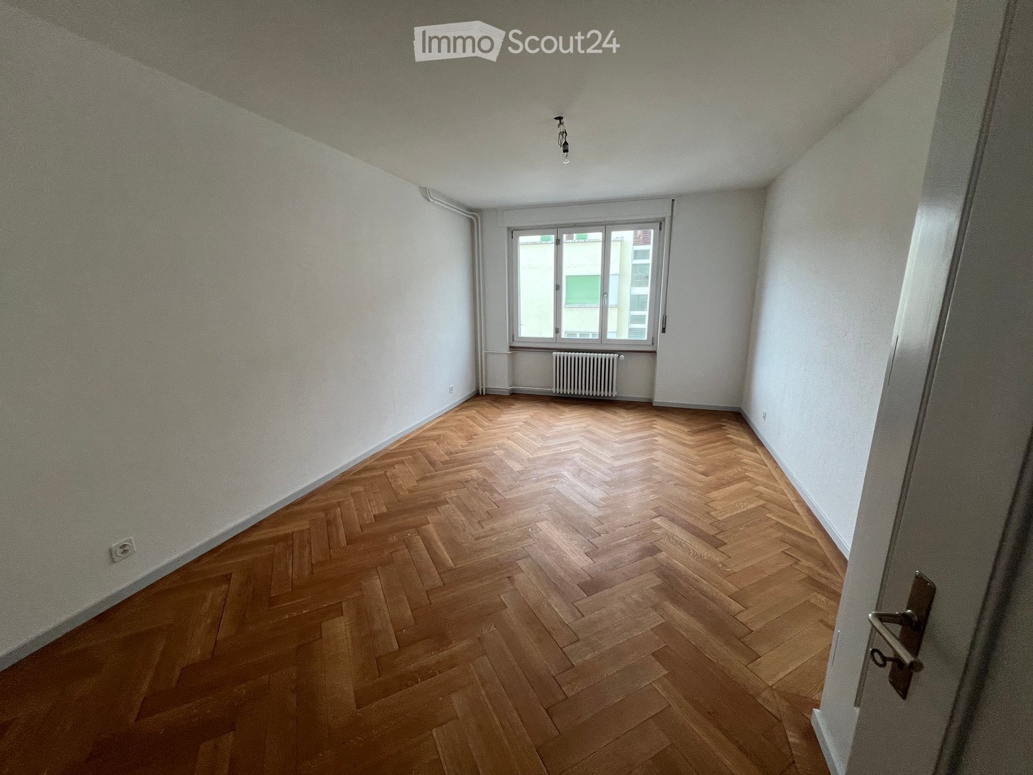 Empty room with wooden herringbone flooring, white walls, and a single window with a radiator below it.