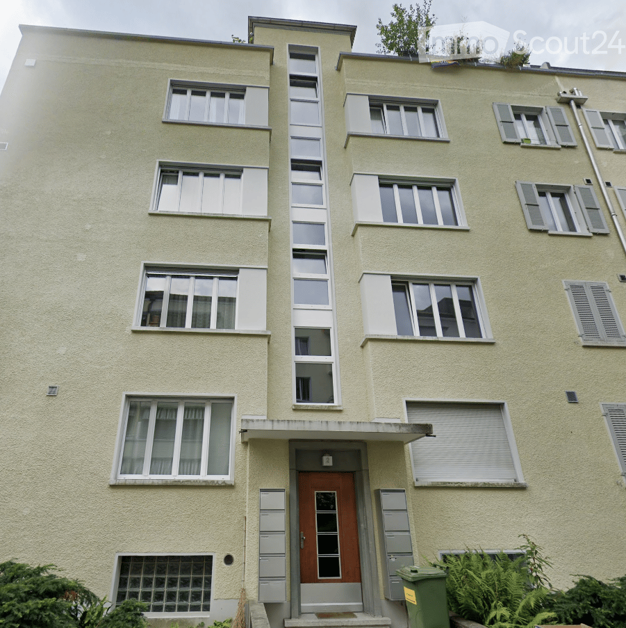 Building. Four-story residential building with light yellow facade, multiple windows, and a central entrance with a small canopy. The building features a mix of rectangular and square windows, some with shutters, and a small garden area in front.