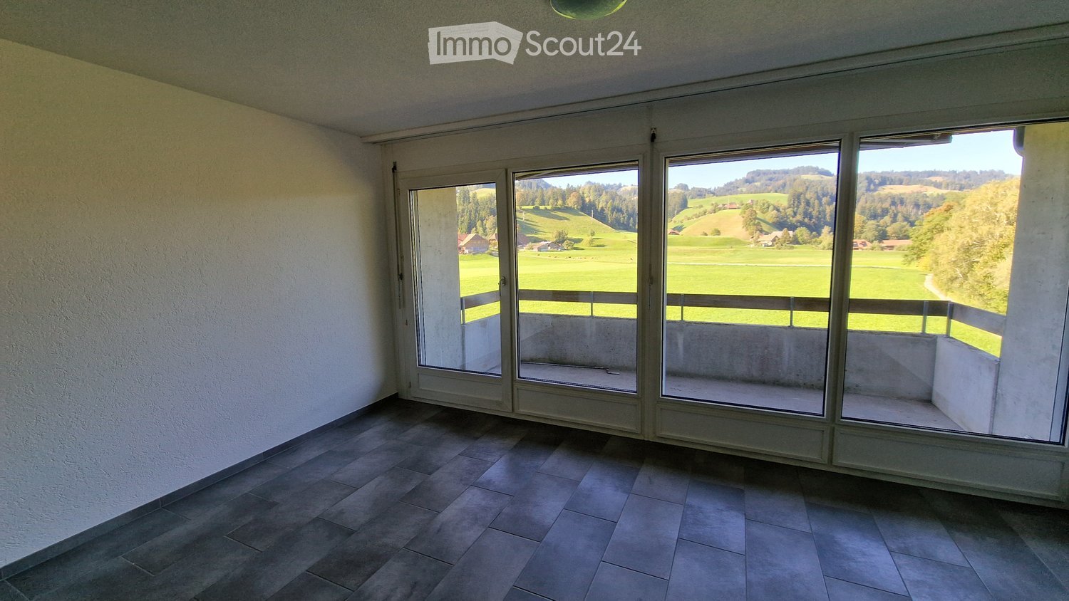 Empty room with large windows offering a scenic view of green fields and hills. The room has tiled flooring and white walls.