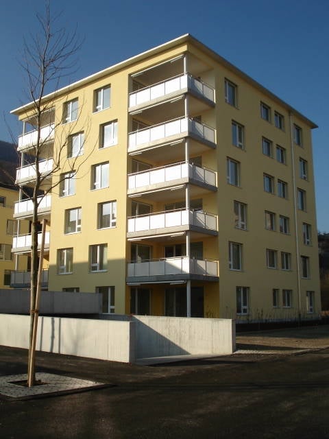Building. A modern yellow building with multiple floors and balconies. The building has a concrete wall and a tree in front.