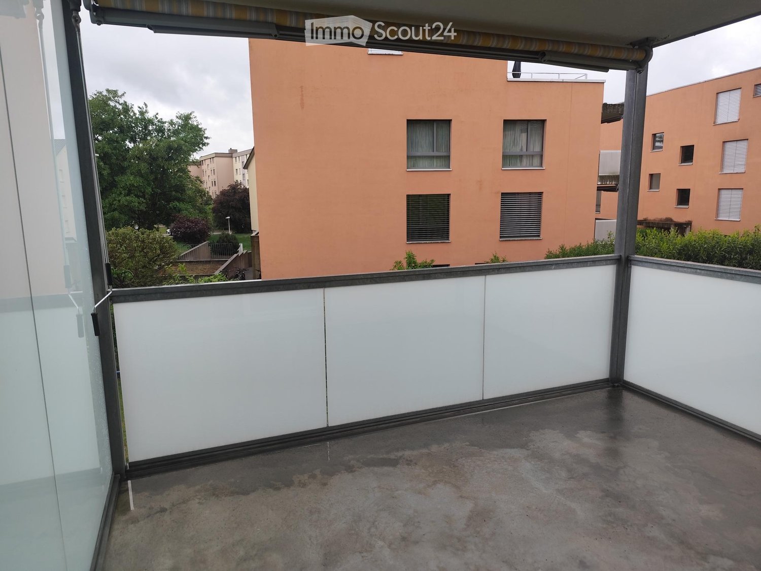 Balcony with white walls, concrete floor, and a view of a residential building and greenery.