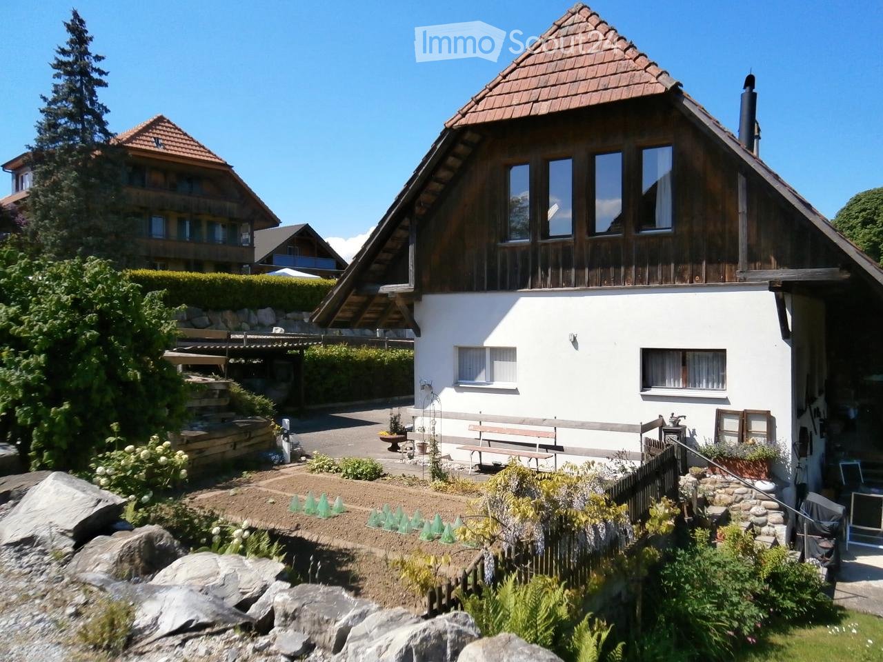 House. A two-story house with a wooden roof and white walls, surrounded by a garden with benches, potted plants, and a small staircase leading to the entrance.