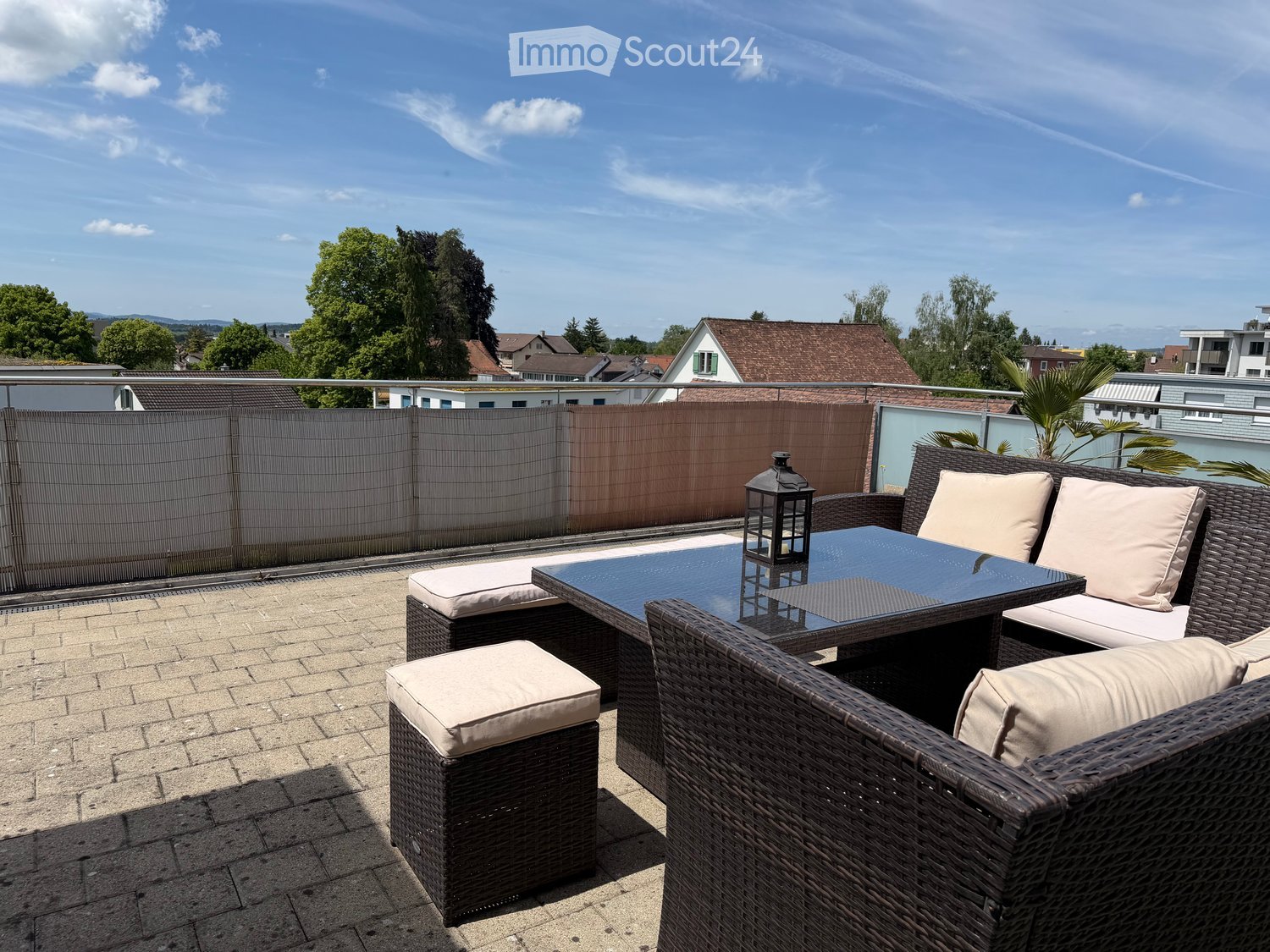 A modern rooftop terrace with a wicker seating set, glass table, and lantern, overlooking a residential area with trees and houses.