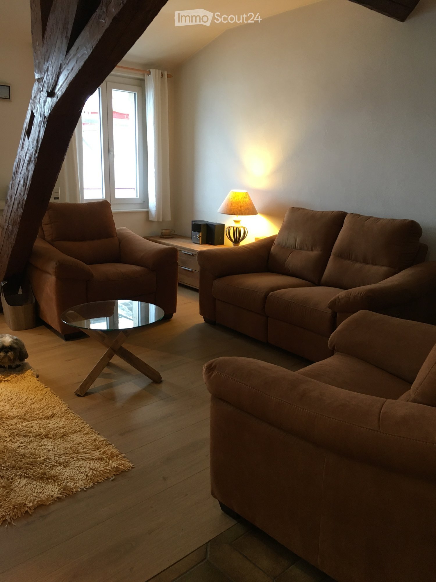 Living room with wooden flooring, large window with white curtains, brown sofa, armchair, glass coffee table, table lamp, side table, and speakers.