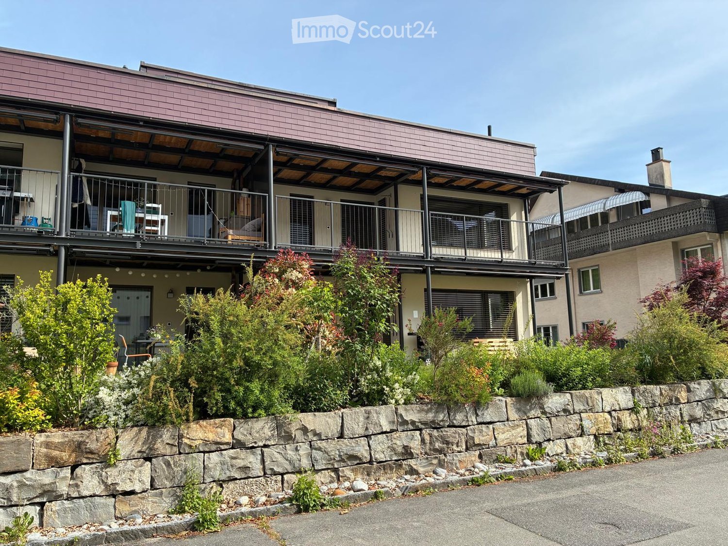 Two-story residential building with a modern design, featuring a covered balcony area, metal railings, and a stone retaining wall with greenery in front.