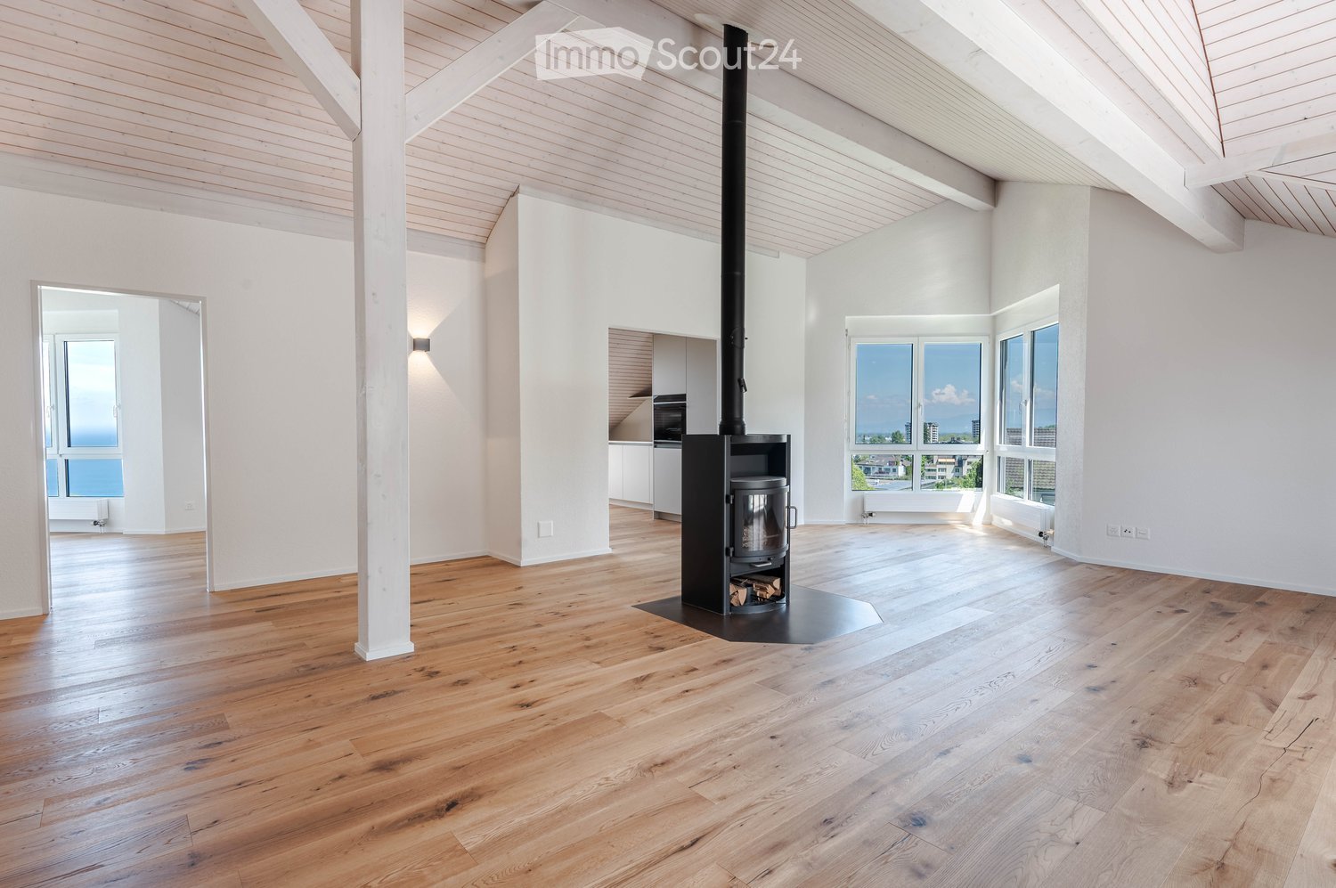 Bright, open living space with wooden flooring, white walls, and large windows. Features a wood stove in the center, a kitchen area with cabinets and an oven, and ample natural light.