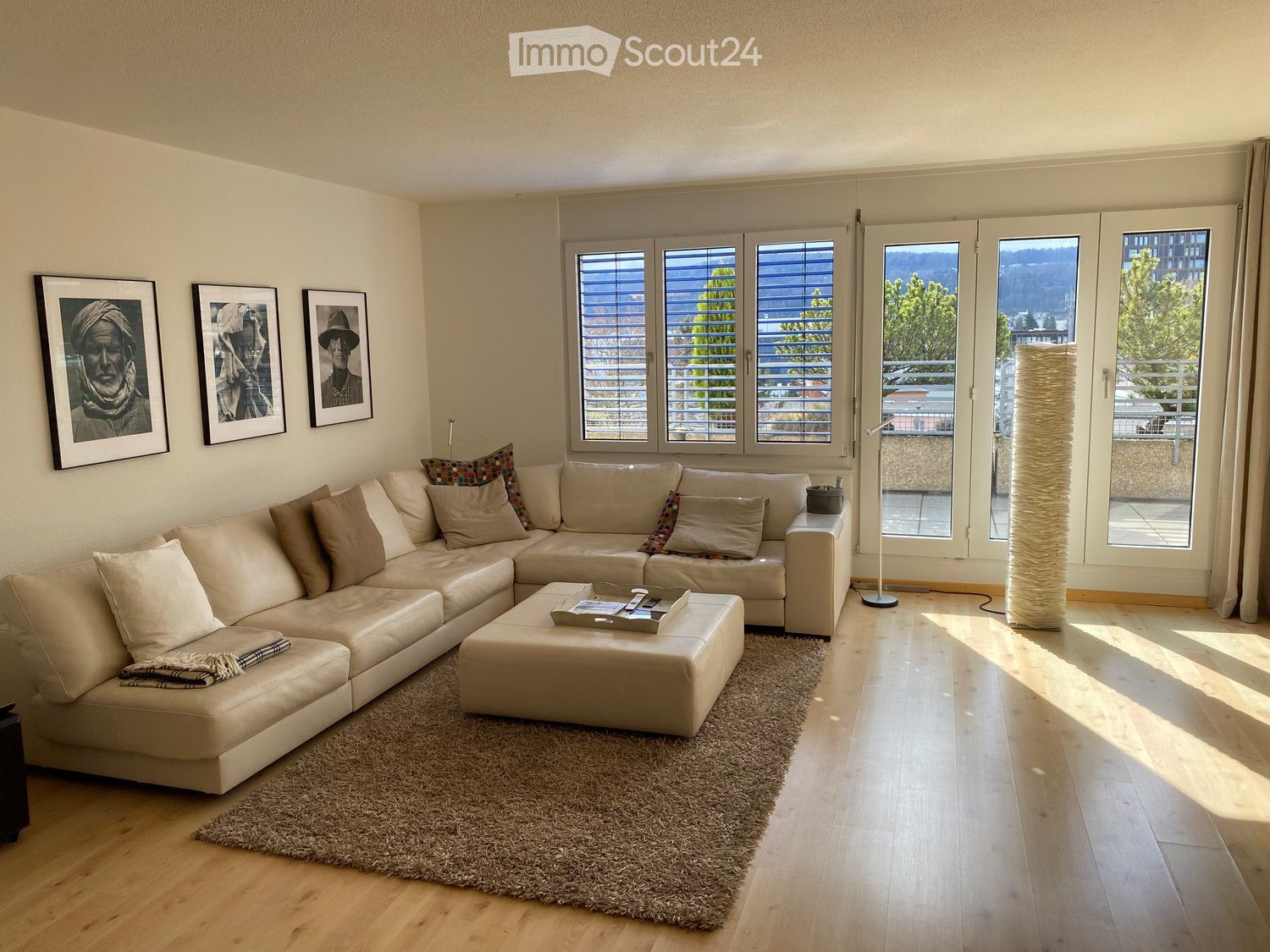 Modern living room with a large sectional sofa, coffee table, ottoman, floor lamp, and artwork on the wall. Large windows offer a scenic view.