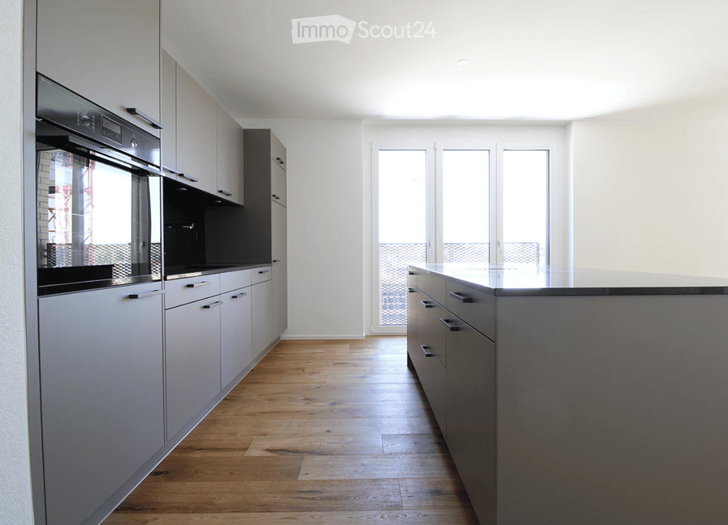 Modern kitchen with grey cabinets, wooden flooring, and an integrated oven. Large windows provide natural light.