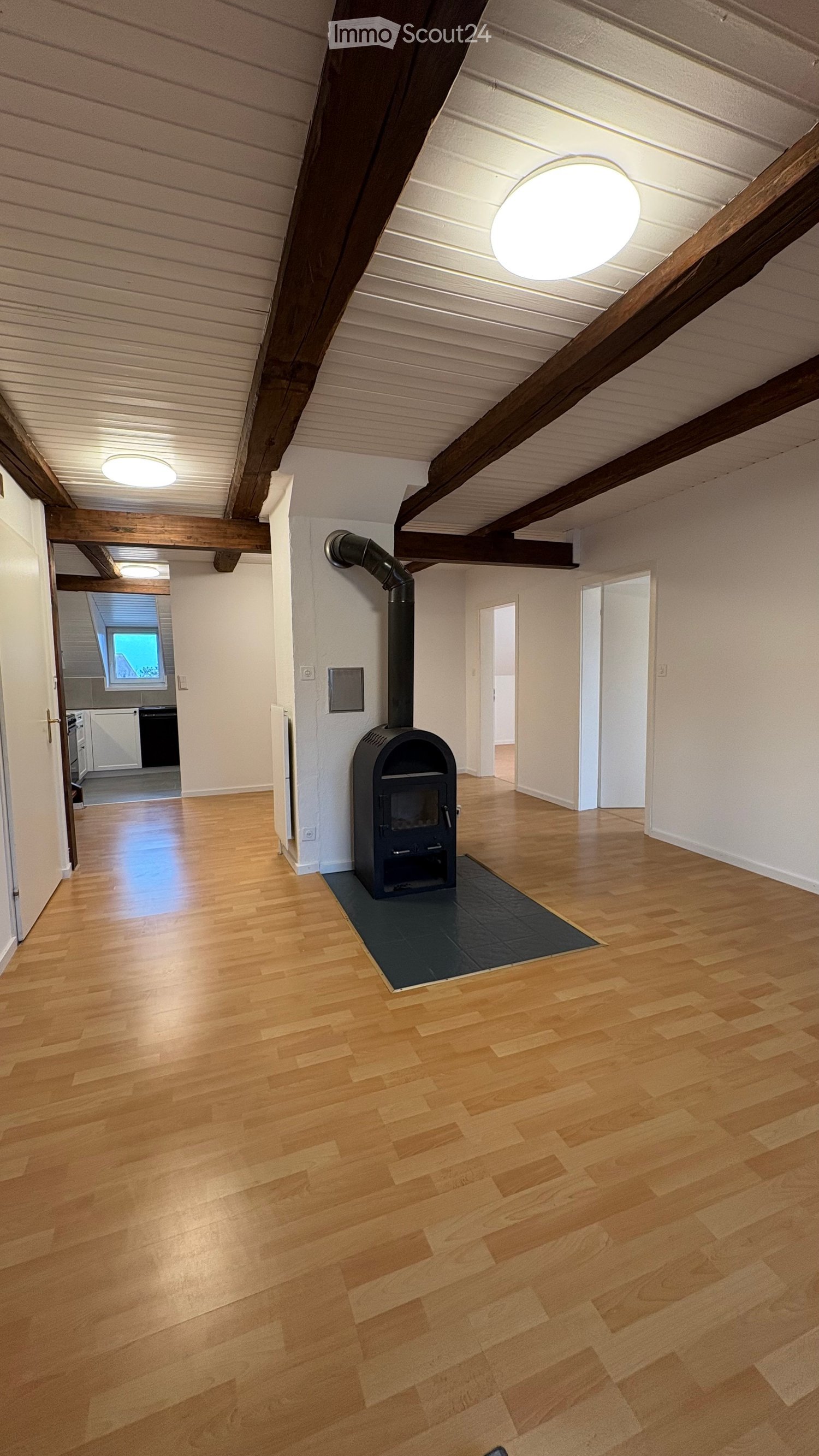 Empty room with wooden flooring, exposed wooden beams, and a black wood-burning stove with a chimney pipe.
