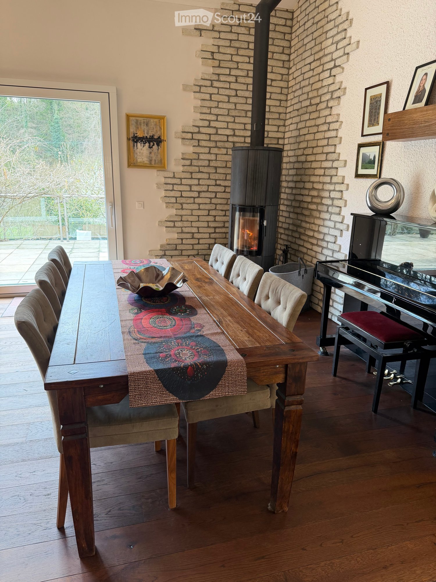 Dining room with wooden floor, fireplace, dining table with chairs, piano, and artwork on the walls.