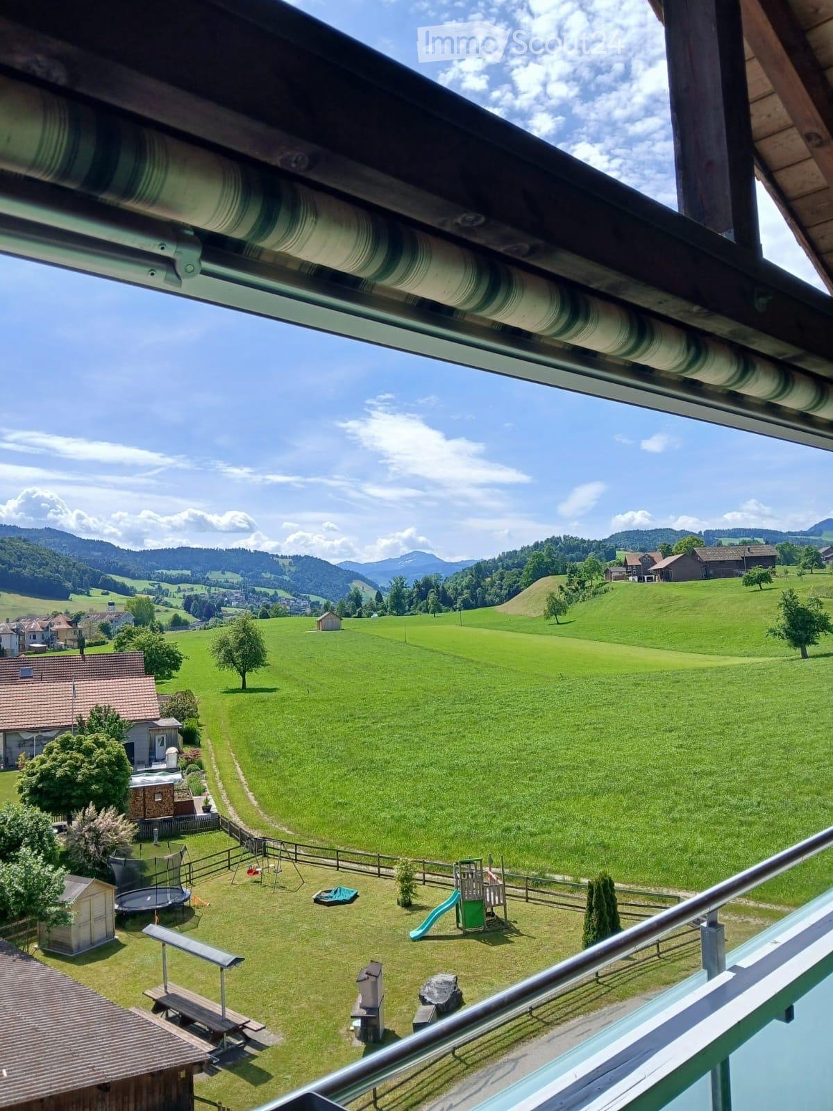 View from a balcony overlooking a spacious, green lawn with a fenced play area, trampoline, swing set, and slide. The background features rolling hills, scattered houses, and distant mountains under a partly cloudy sky.