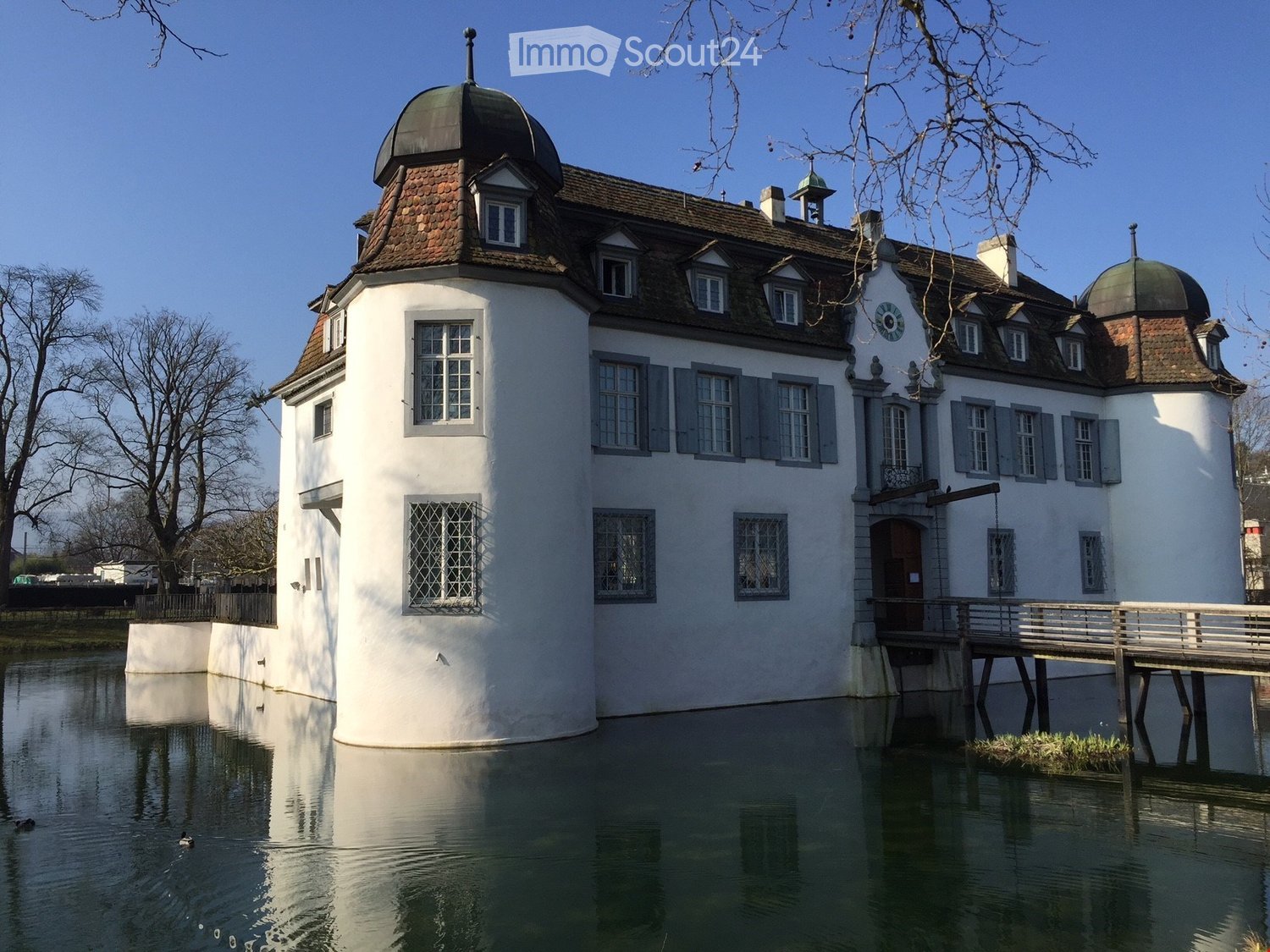 A large, white, historic building with a dome and multiple windows, situated beside a calm body of water with a wooden bridge leading to it.