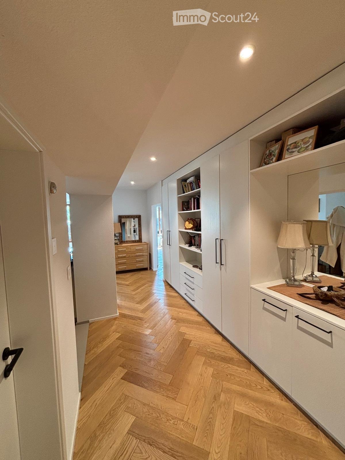 A well-lit hallway with wooden flooring, white built-in storage units, and a small table with lamps and decorative items.