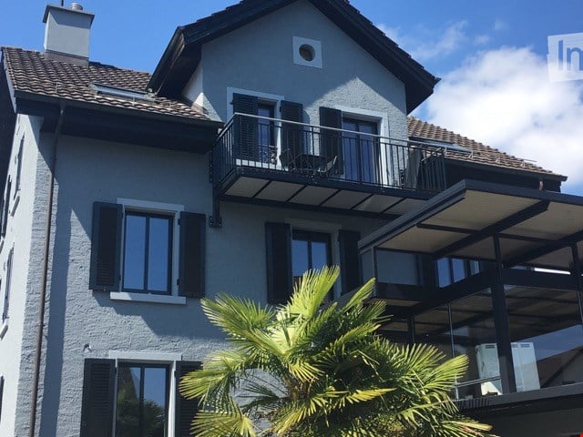 Two-story house with light gray exterior, dark shutters, and a balcony with metal railings. The roof is tiled, and there is a small chimney on the left side.