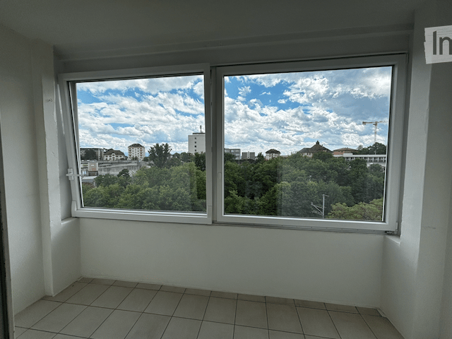 Empty room with large windows offering a view of trees and buildings. The room has tiled flooring and white walls.