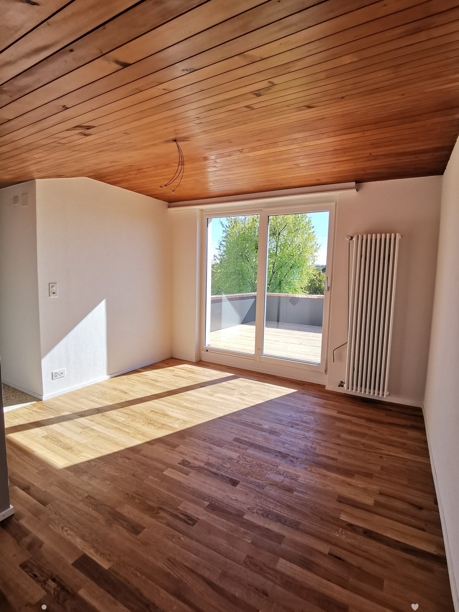 Empty room with wooden flooring, white walls, and large windows leading to a balcony. The ceiling is also wooden, and there is a radiator on one wall.
