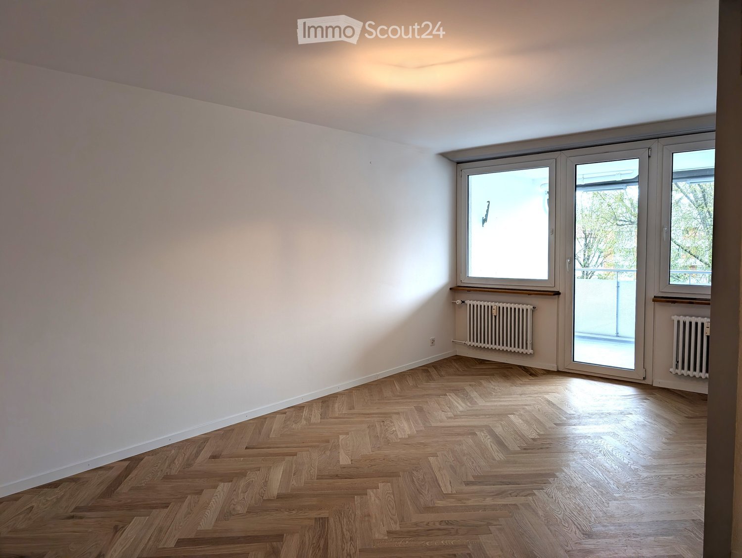 Empty room with wooden herringbone flooring, white walls, and large windows with sliding doors leading to a balcony.