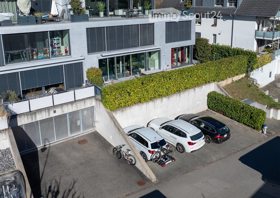 Modern multi-story residential building with large windows, balconies, and a parking area with several cars and bicycles.