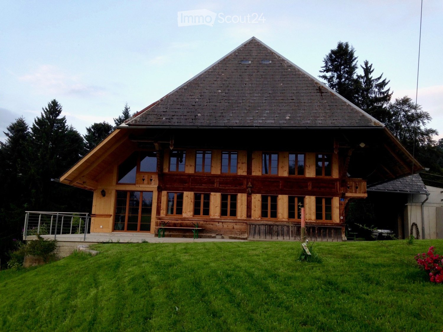 House. Two-story wooden house with a steep roof, large windows, and a raised ground floor. The house features a wooden balcony and is surrounded by a well-maintained lawn with trees and flowers.
