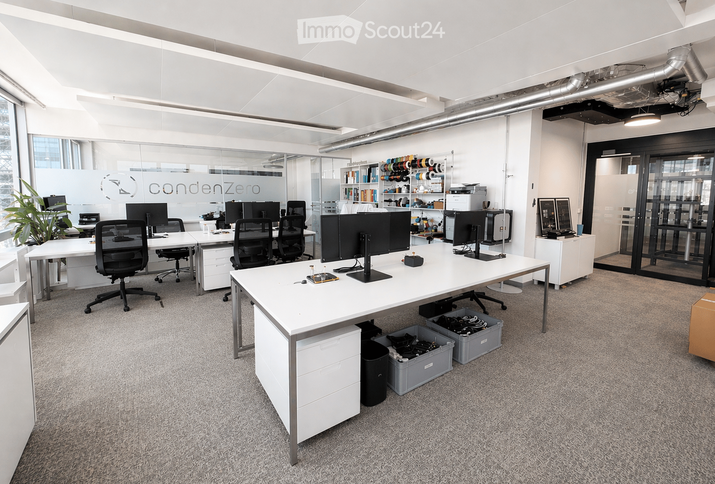 Modern office space with white desks, black office chairs, multiple computer monitors, shelves with office supplies, and a potted plant near the window.