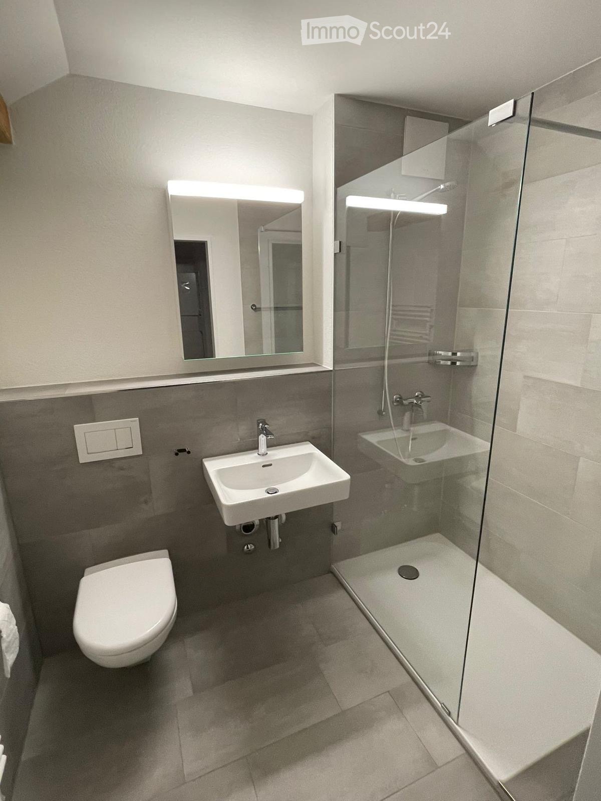 Modern bathroom with a toilet, sink, mirror, and glass-enclosed shower. Grey tiled walls and floor, white fixtures.