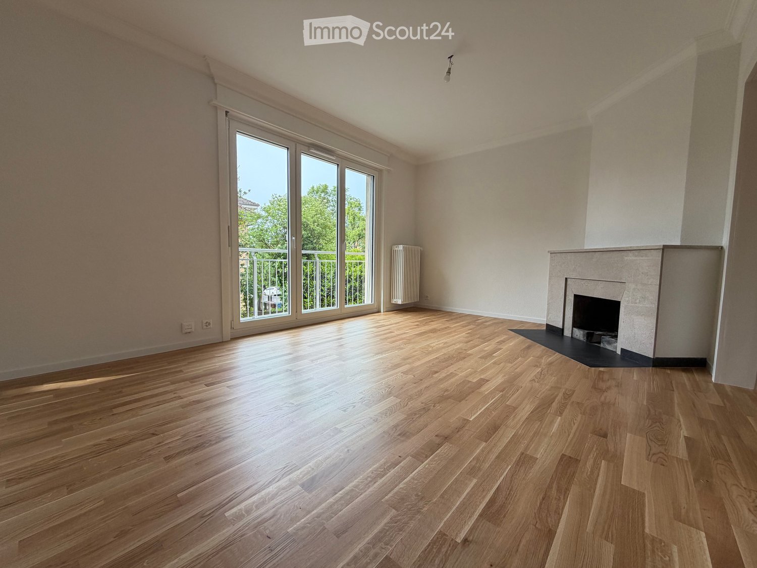 Empty room with light wooden flooring, white walls, large sliding glass doors leading to a balcony, and a fireplace with a white surround.