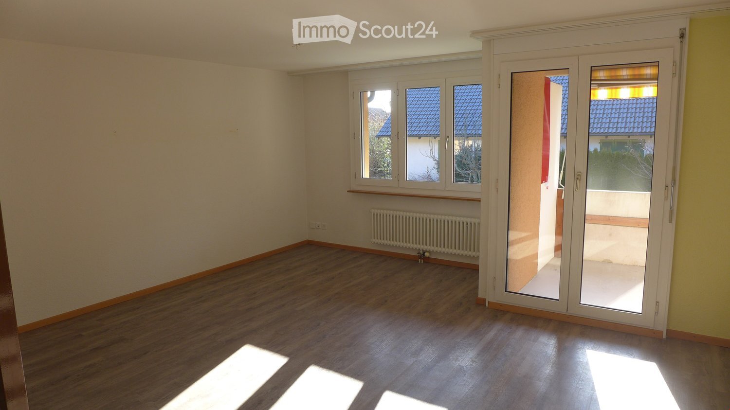 Empty room with wooden flooring, white walls, and large windows with a sliding glass door leading to a balcony.