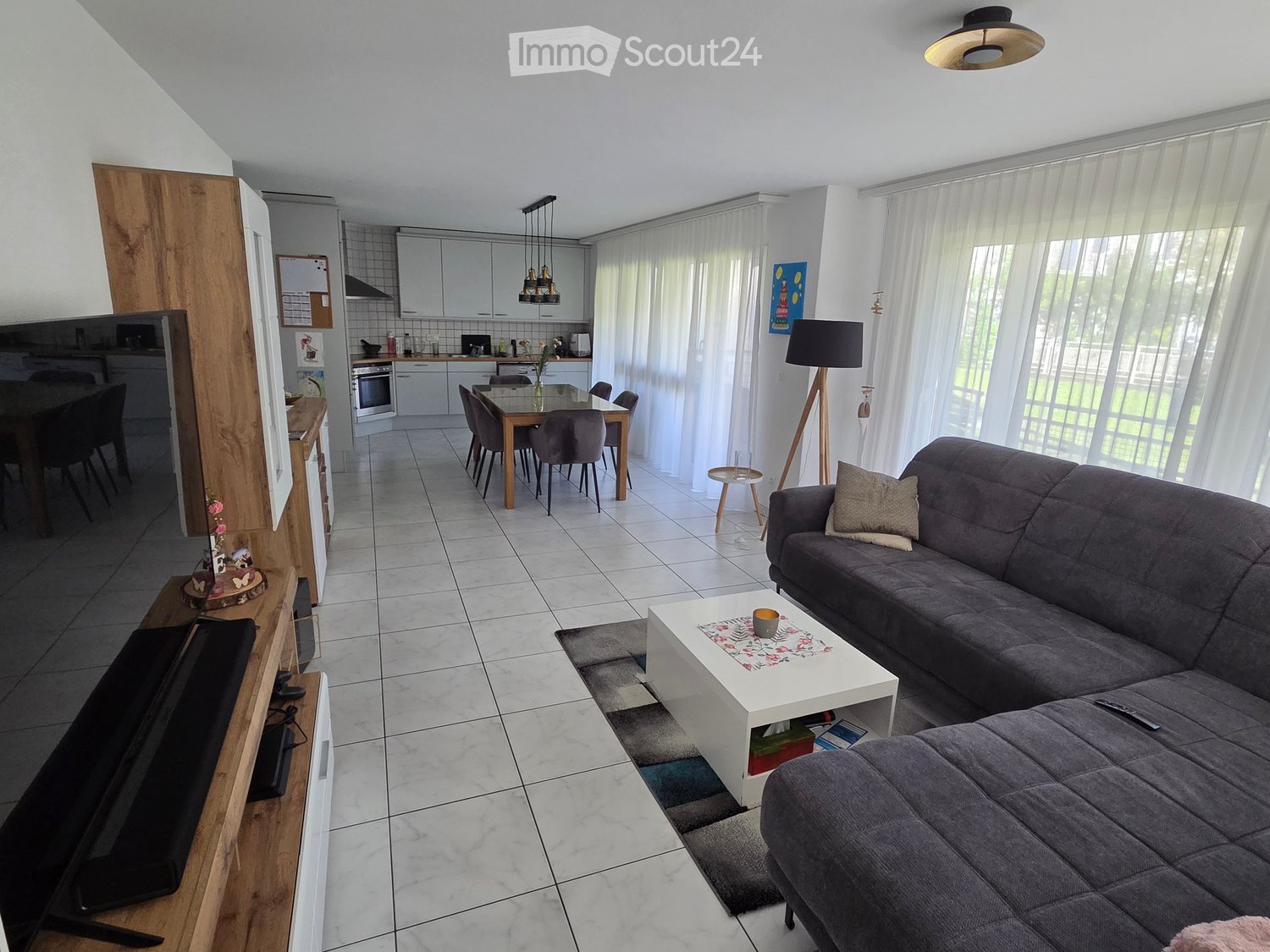 Open-plan living and dining area with a grey sectional sofa, white coffee table, dining table with chairs, and a modern kitchen in the background.