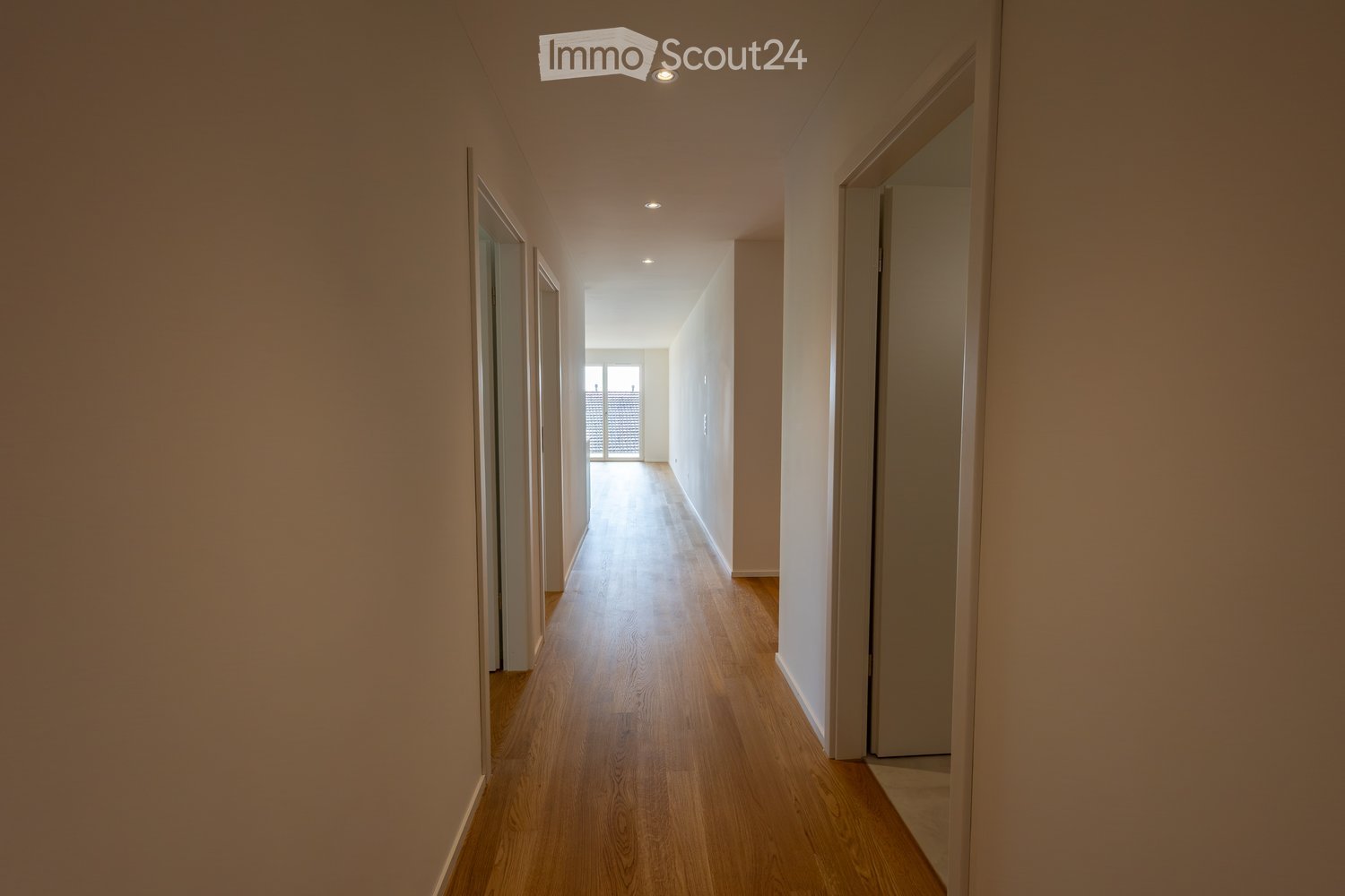 A long, narrow hallway with light wooden flooring and white walls, featuring recessed ceiling lights and doors on both sides leading to other rooms.