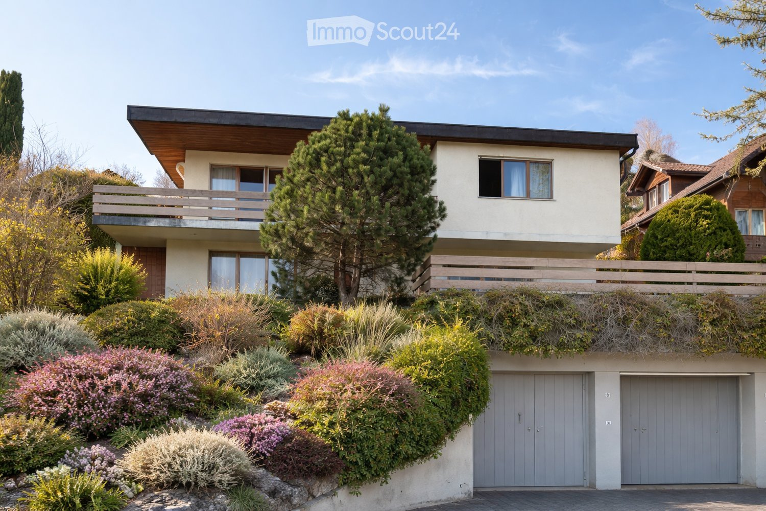 Two-story house with a raised ground floor, modern design, large windows, and a balcony on the upper level. The house features a garage and is surrounded by well-maintained gardens and trees.