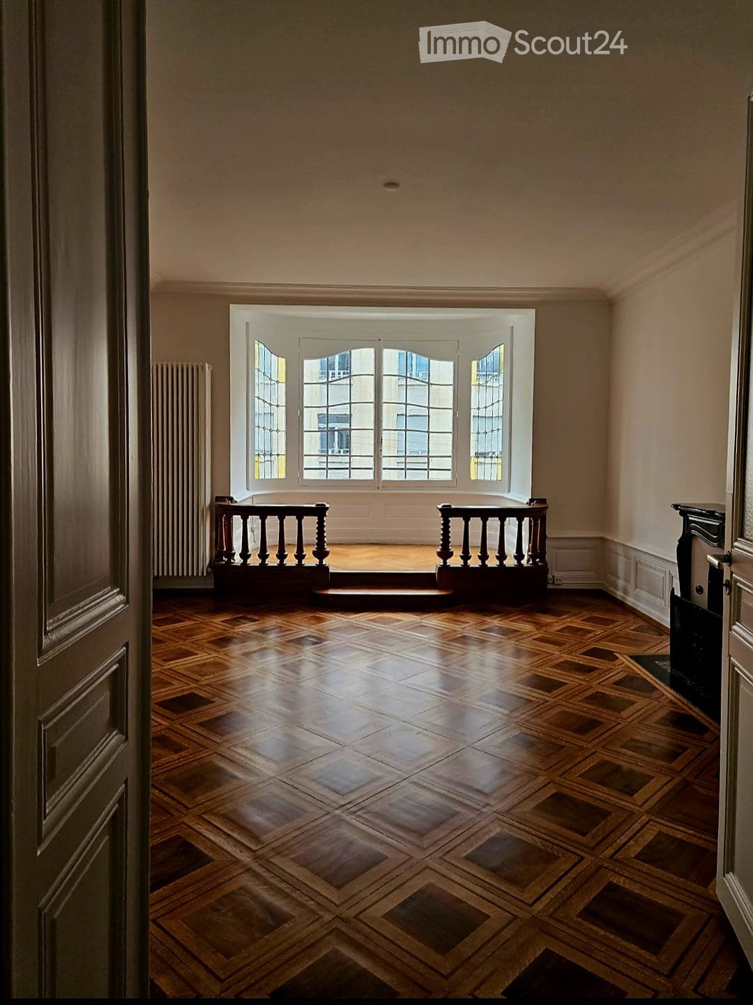 An empty room with wooden herringbone flooring, large windows with white frames, and a small balcony area with a wooden railing.