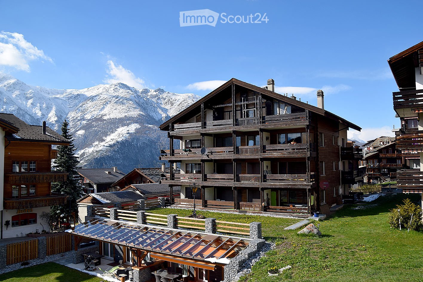 A multi-story wooden building with balconies and a spacious lawn in front, surrounded by snow-capped mountains and other structures.