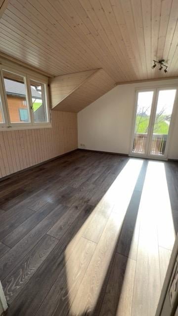 Bright, empty attic room with wooden ceiling, dark hardwood floor, large windows, and a sliding glass door leading to a balcony.