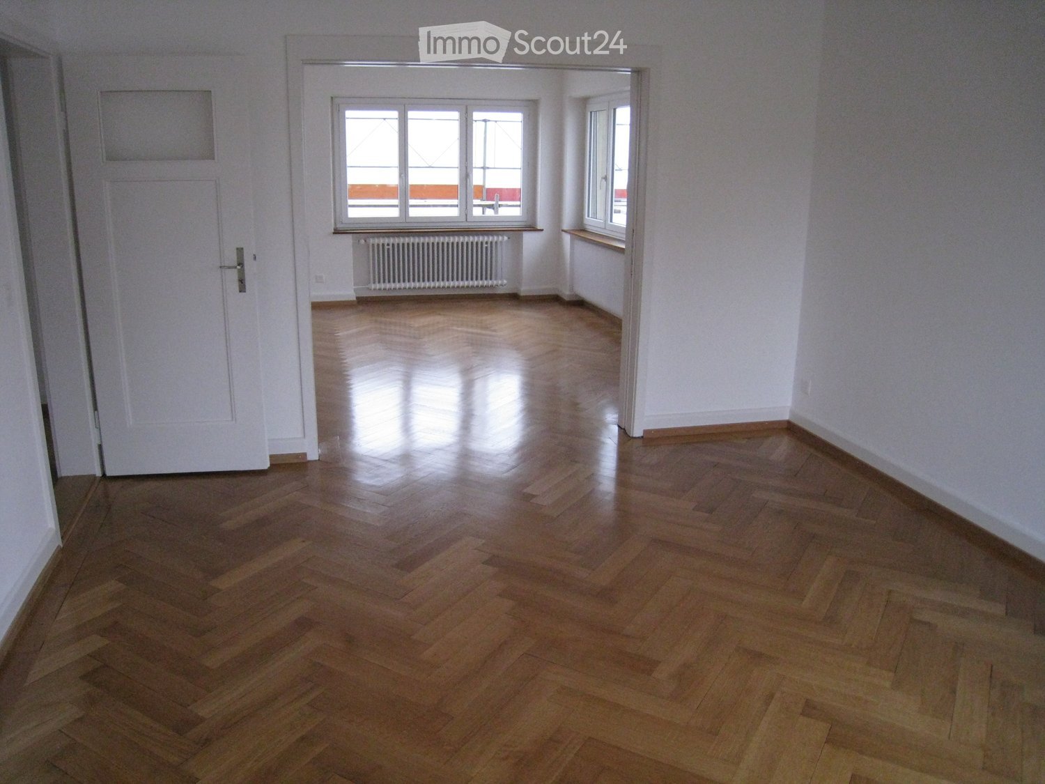 Unfurnished Room. Empty room with wooden flooring, white walls, and large windows allowing natural light. A radiator is visible under one window, and a door is on the left side.