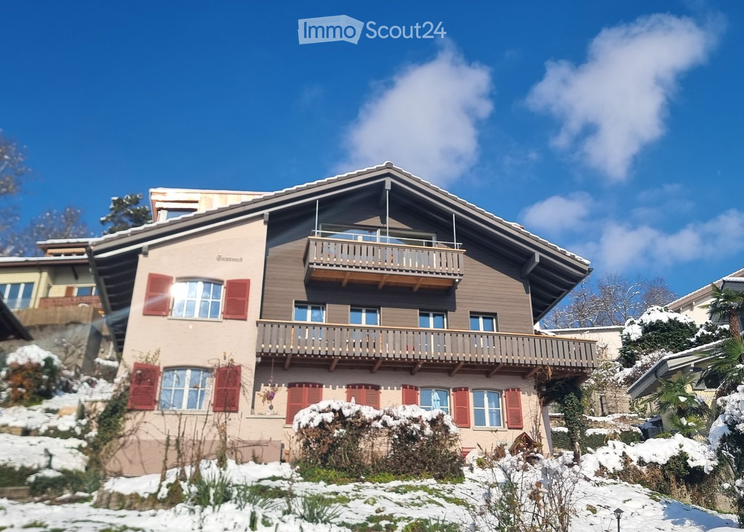 A two-story house with a sloped roof, multiple windows with red shutters, and balconies overlooking a snowy landscape.