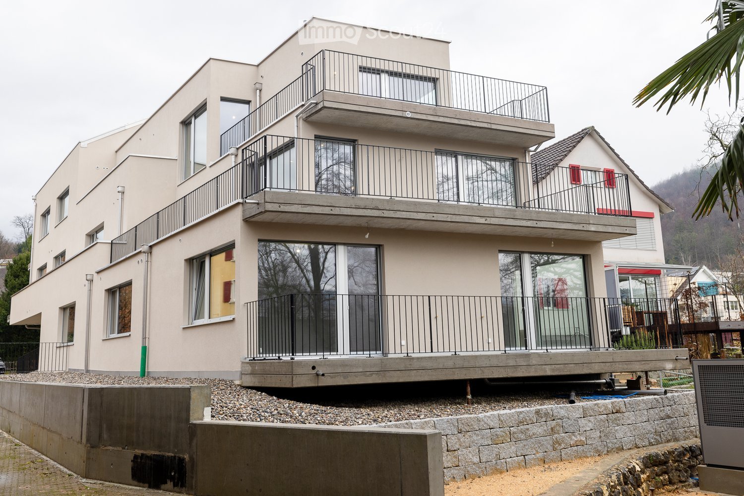 Modern two-story house with beige facade, large windows, multiple balconies, and a raised ground floor.