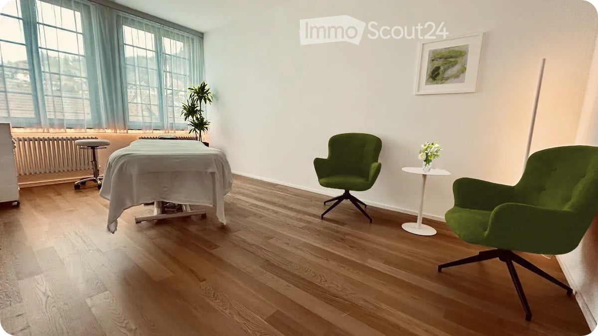 A well-lit room with wooden flooring, a massage table covered with a white sheet, two green chairs, a small round table with a vase of flowers, and a potted plant near the window.