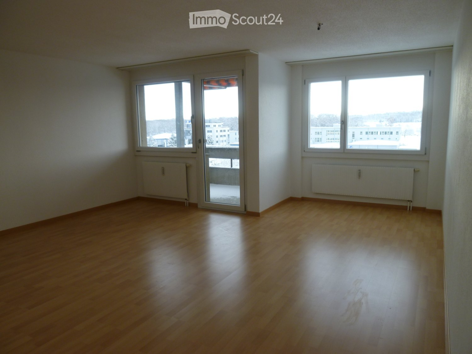 Empty room with wooden flooring, white walls, large windows, and a sliding glass door leading to a balcony.