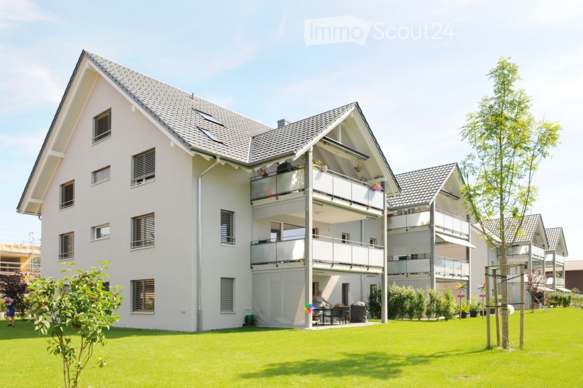 Modern two-story house with white exterior, multiple windows, and balconies. The house is surrounded by a well-maintained lawn and trees.